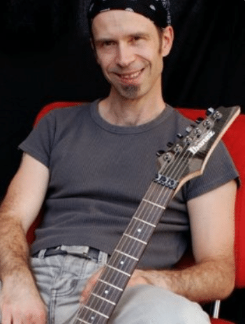 Andreas Vockrodt entspannt mit E-Gitarre, grauer T-Shirt, rote Stuhllehne im Hintergrund