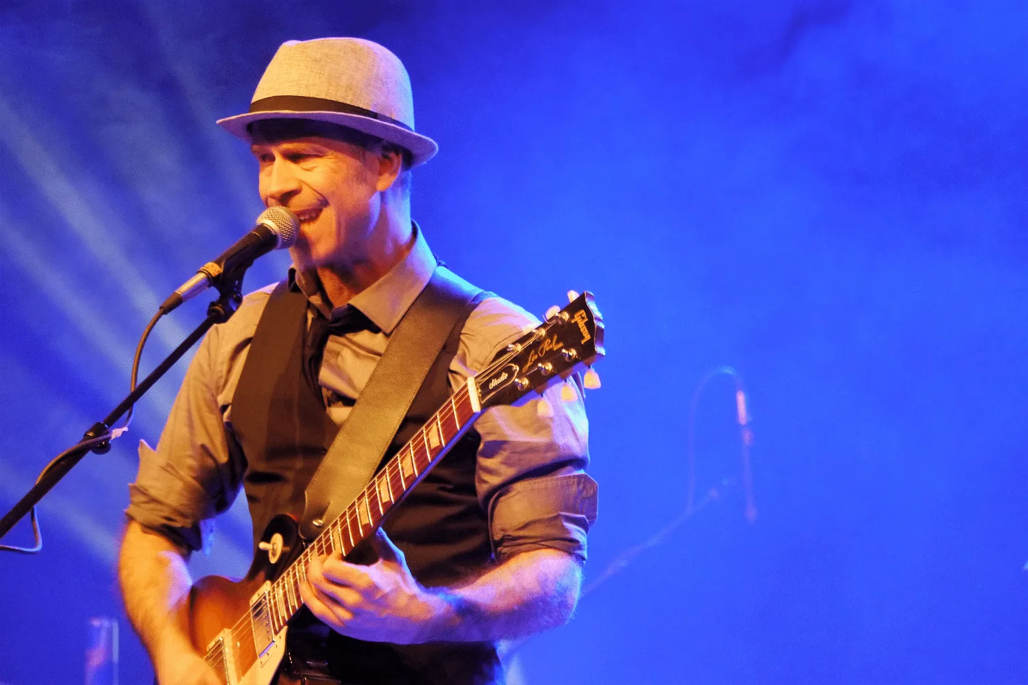 Andreas Vockrodt auf Bühne mit E-Gitarre, Hut, Mikrofon, blaue Bühnenbeleuchtung