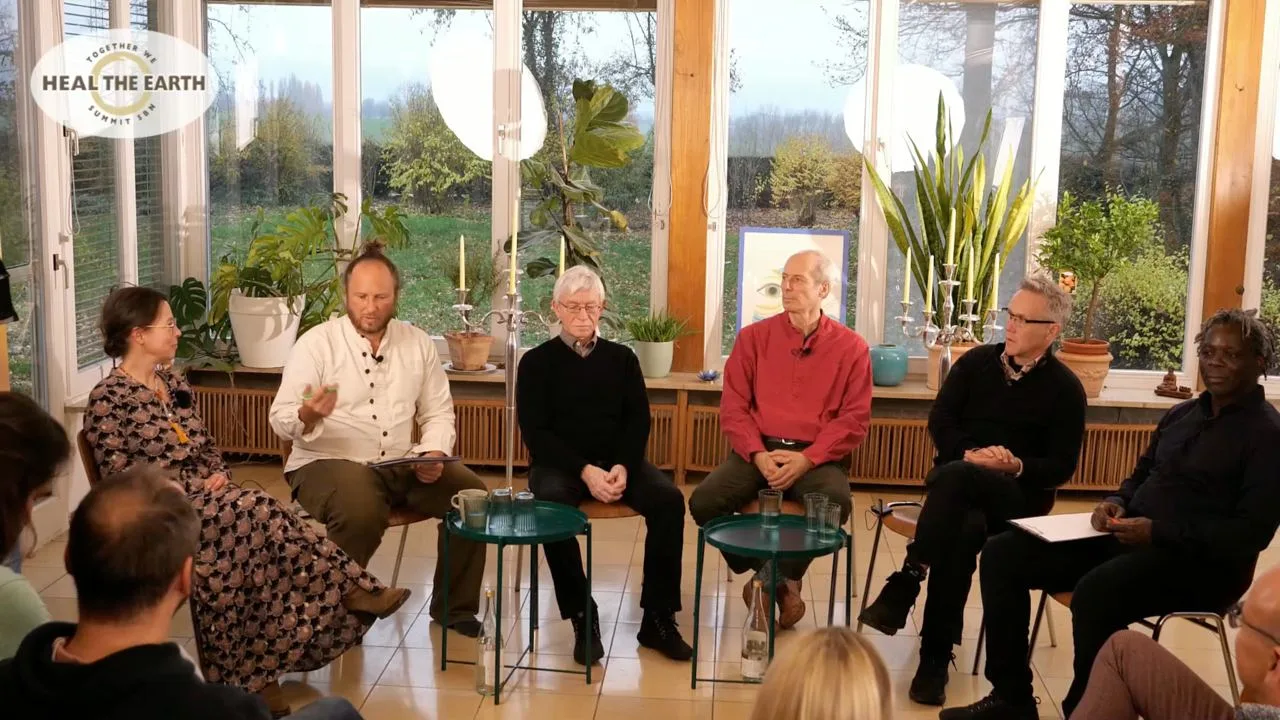 Videostandbild von Diskussionsrunde mit sechs Personen in hellem Raum mit großen Fenstern und Pflanzen