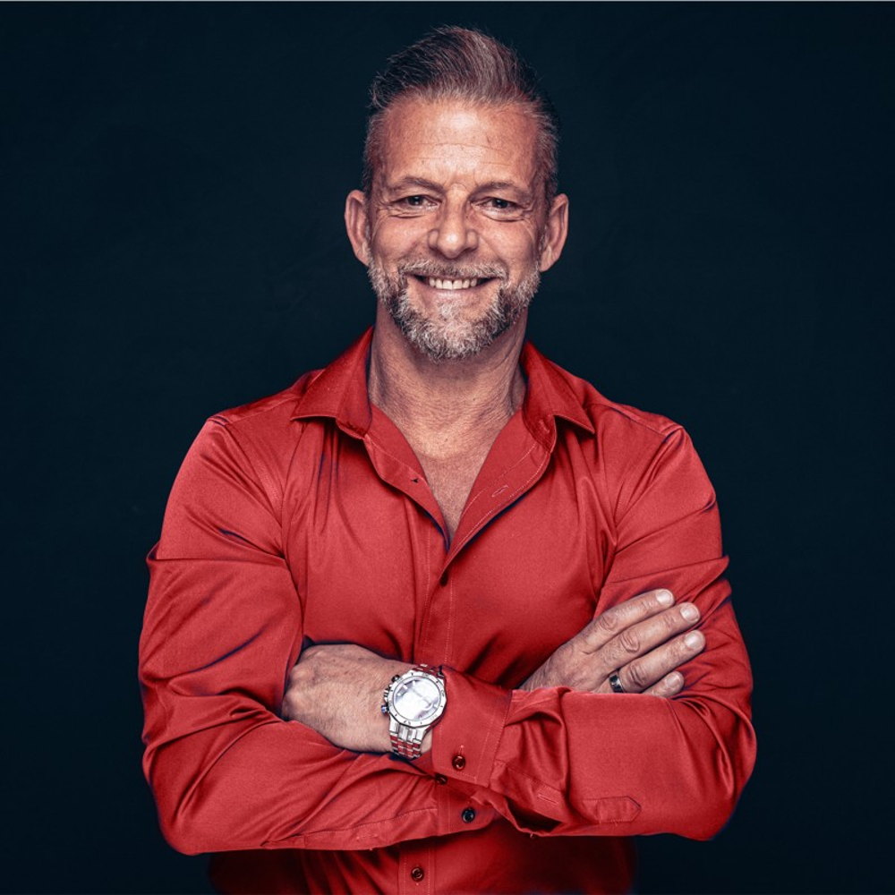 Fabian Kowallik in rotem Hemd vor dunklem Hintergrund, lächelnd mit verschränkten Armen