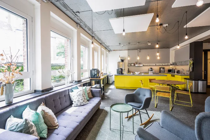 Moderner Arbeits- und Loungebereich mit grauer Polstercouch, gelben Stühlen, Küchenzeile und großzügigen Fensterfronten