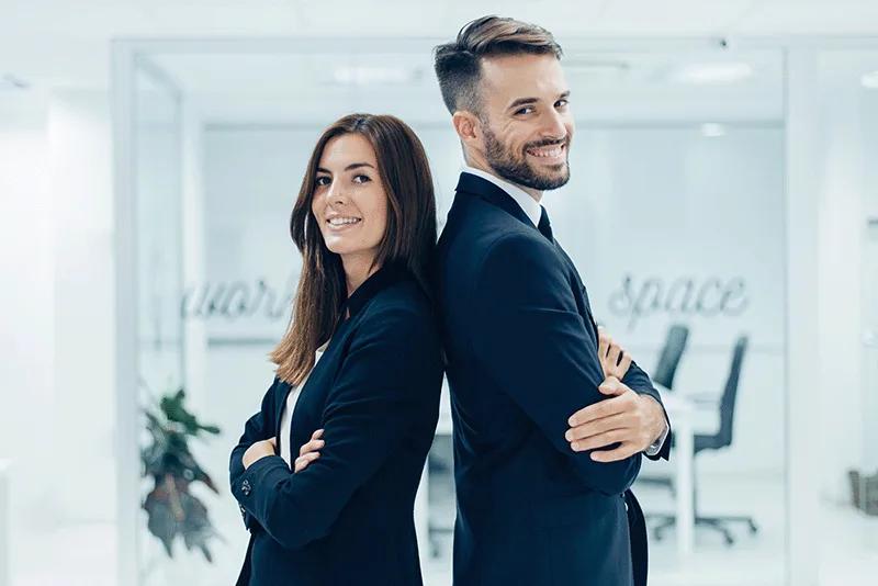 Geschäftsfrau und Geschäftsmann in dunkelblauen Anzügen stehen back-to-back in modernem Büro, beide lächelnd