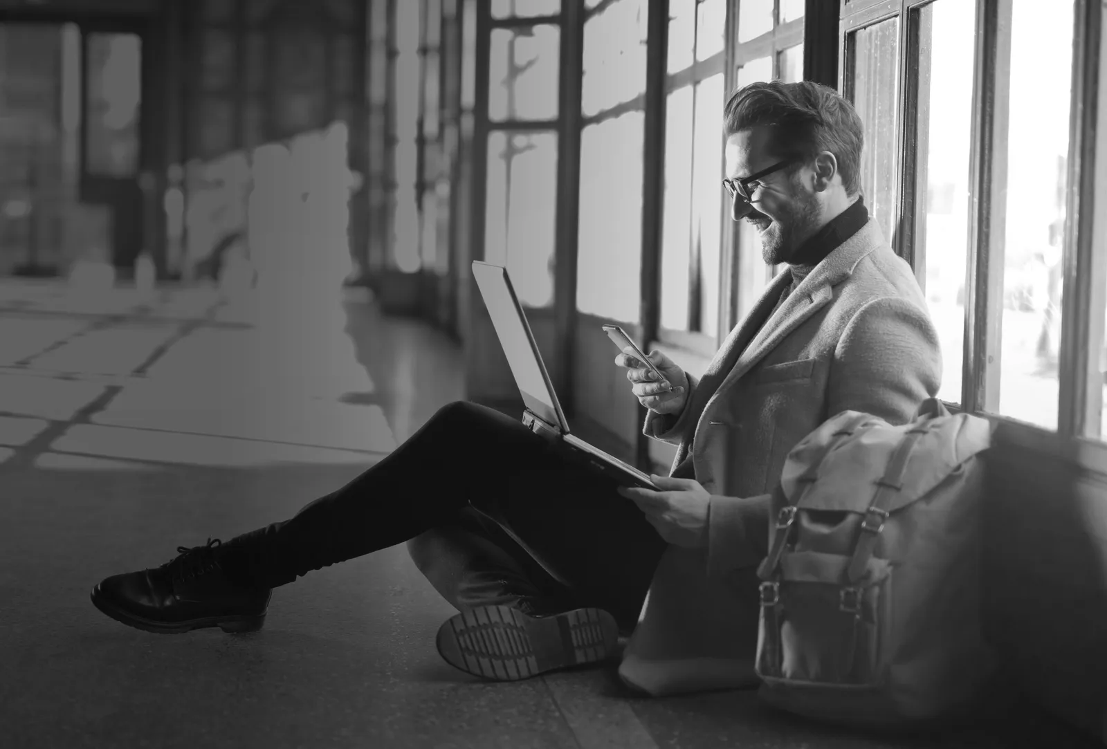 Mann mit Brille sitzt am Fenster mit Laptop und Smartphone, konzentriert arbeitend, Schwarz-Weiß-Fotografie