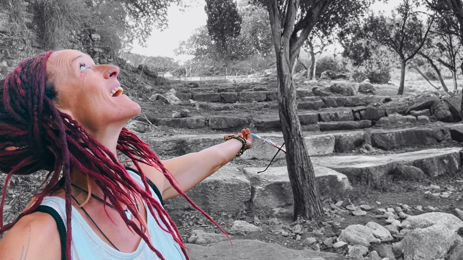 Frau mit roten Dreadlocks lacht, streckt Arm aus vor alten Steinstufen und Wald