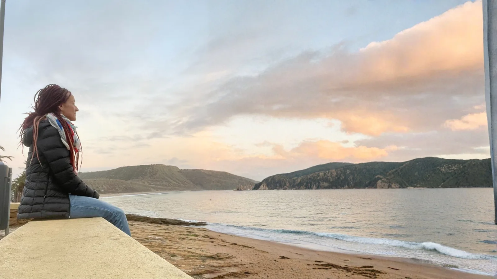 Frau in Winterjacke sitzt an Küste und blickt auf Meer mit Bergen bei Sonnenuntergang