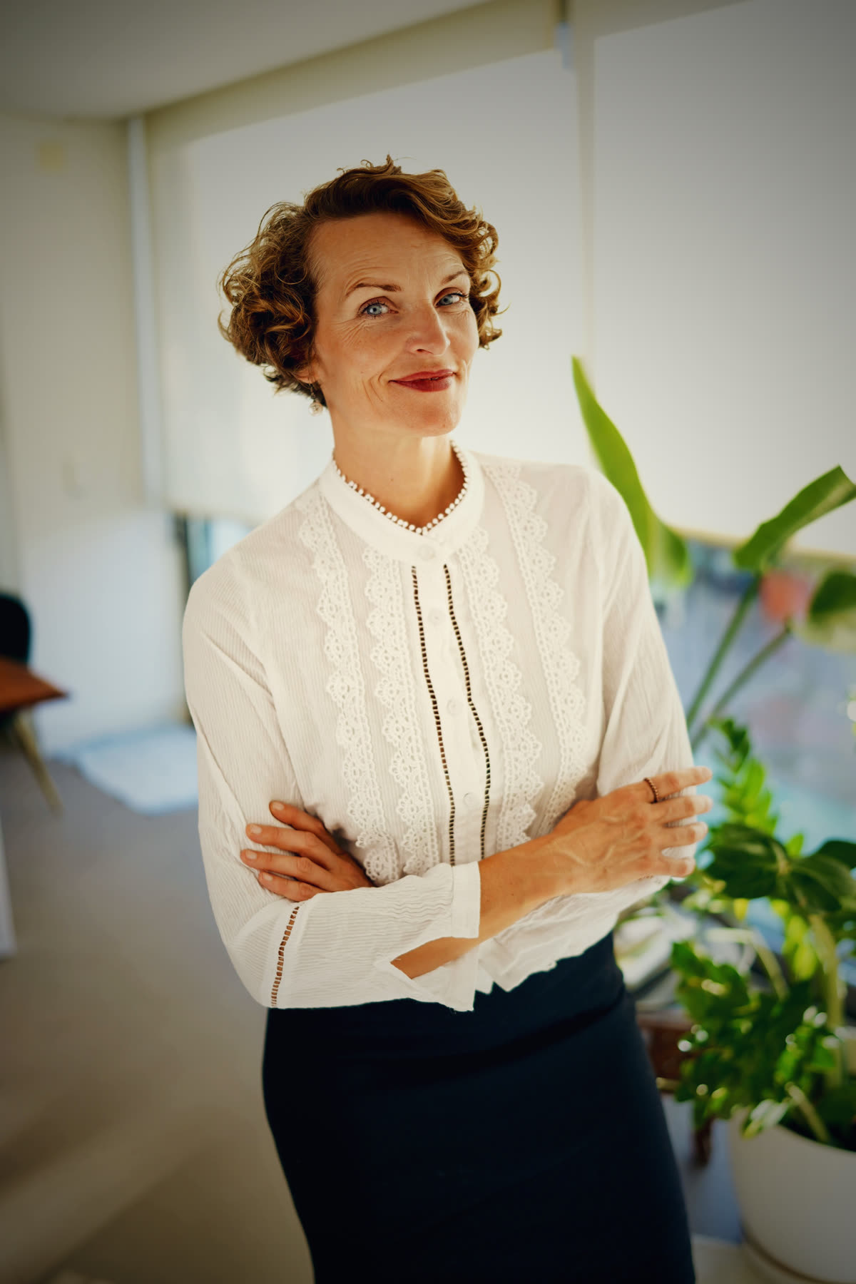Stefanie Bruns in weißer Spitzenjacke mit Perlenkette, verschränkte Arme, moderne Inneneinrichtung