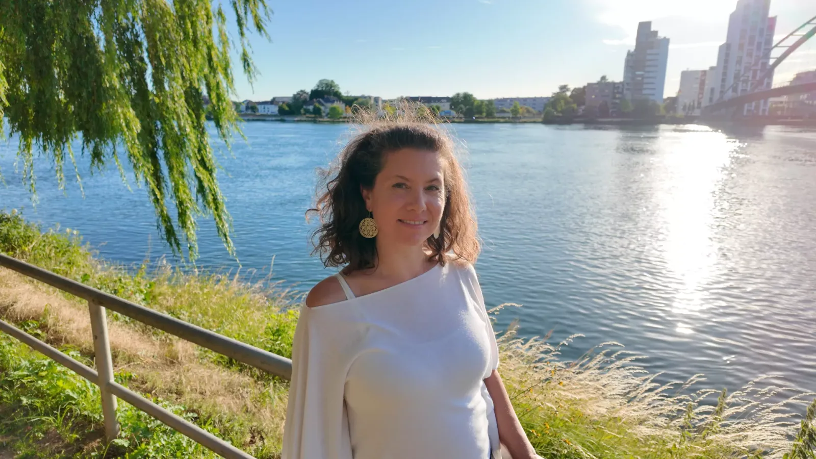 Sabine Aktar am Fluss mit Stadtkulisse, weiße Bluse, Gold-Ohrringe