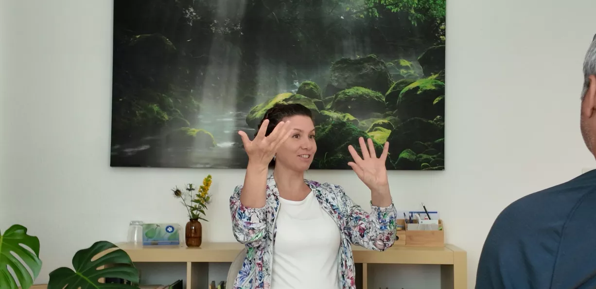 Sabine Aktar in Praxis-Setting, gestikulierend während Beratung, helles Büro mit Waldbach-Bild