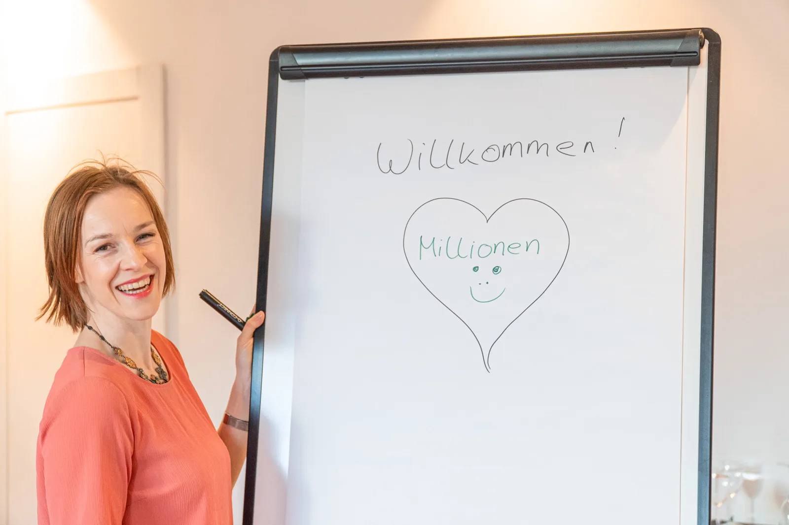 Energetische Heilung Community von Julia Gössler 3 Frau mit rötlichen Haaren in korallfarbenem Shirt hält Whiteboard mit 'Willkommen Millionen' und gezeichnetem Herz-Smiley