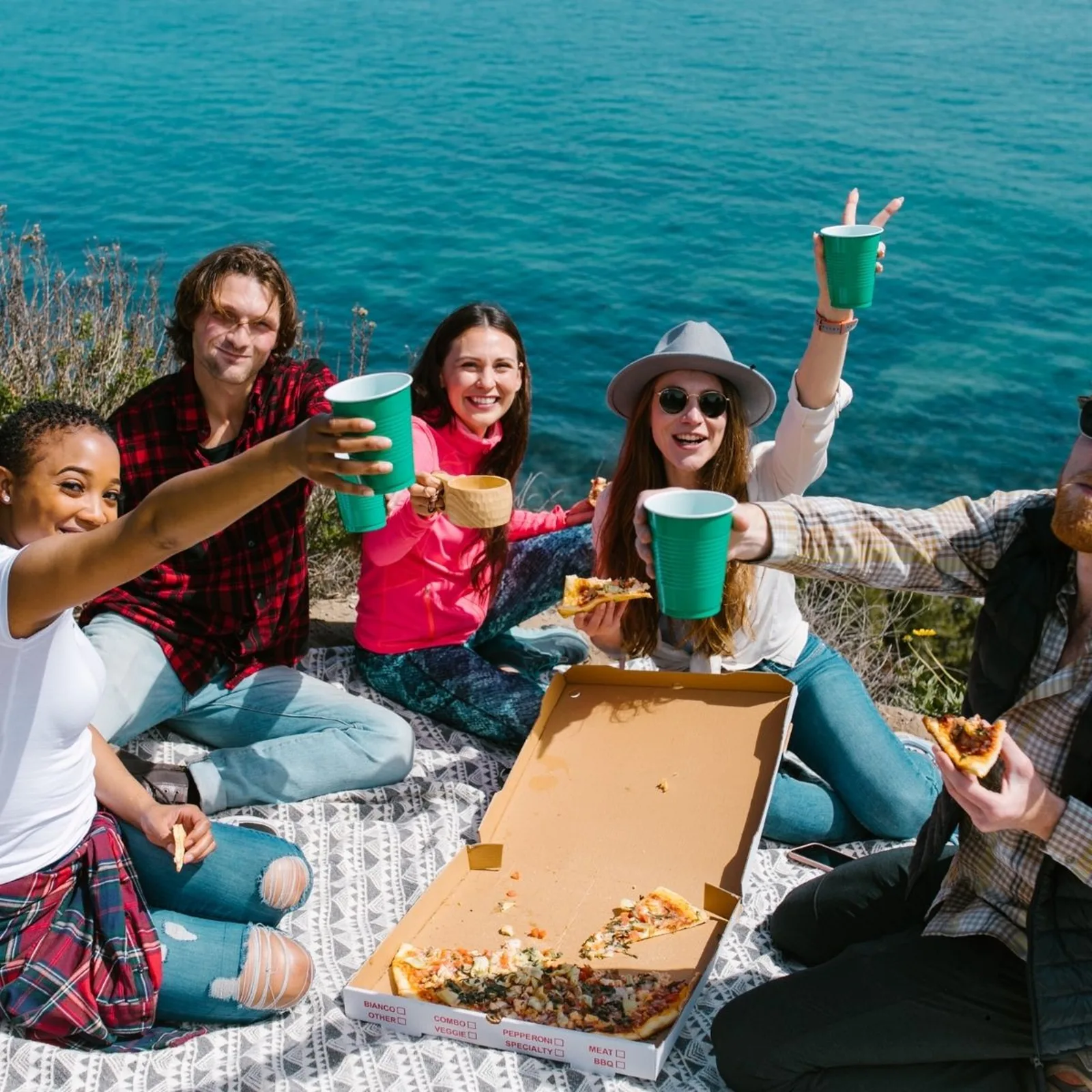 Gruppe von fünf Personen feiert an Meeresküste mit Pizza und grünen Getränken
