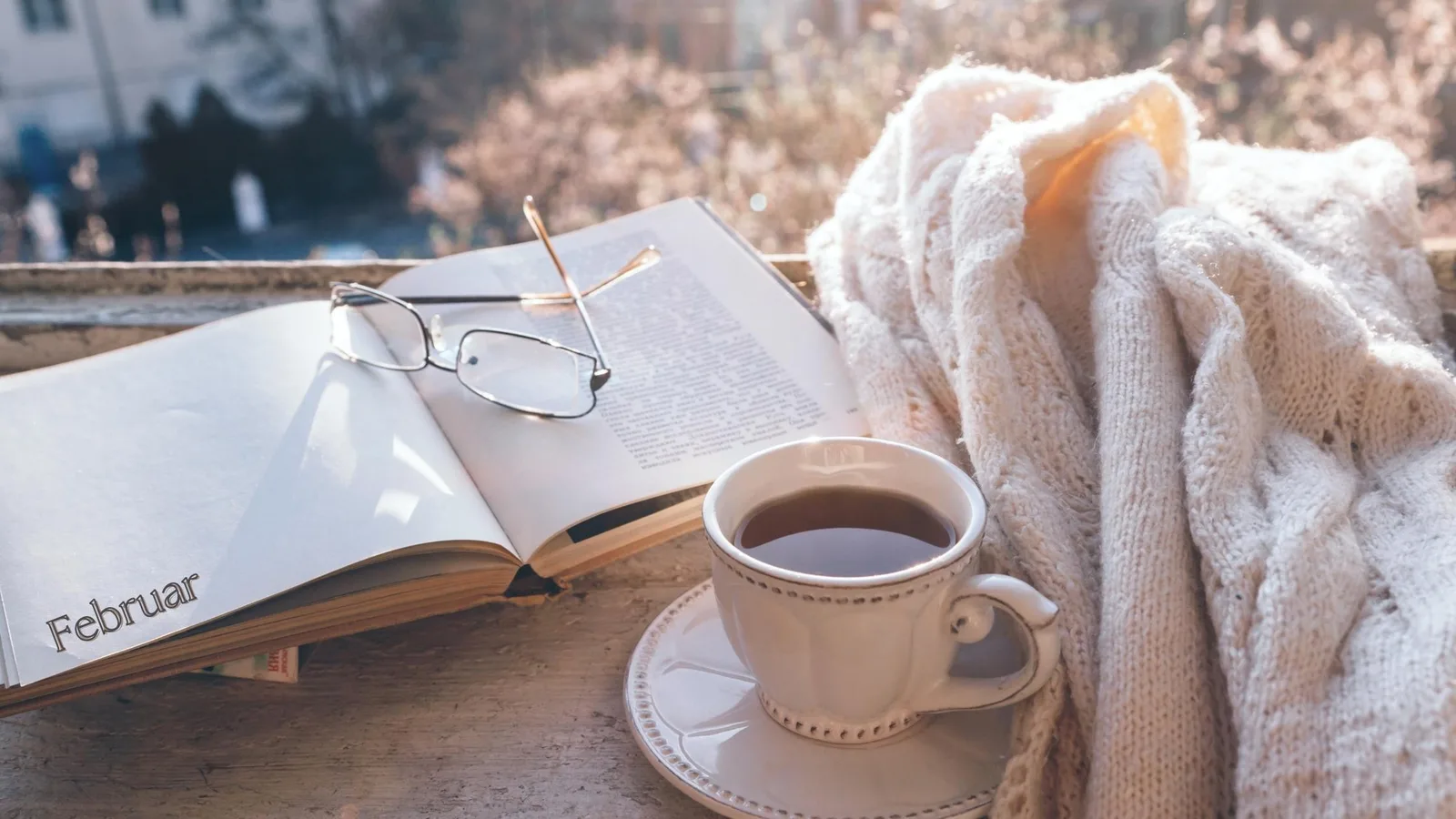 Buch aufgeschlagen mit Februar-Seite, Brille, Kaffee und Schal auf Stein am Fenster