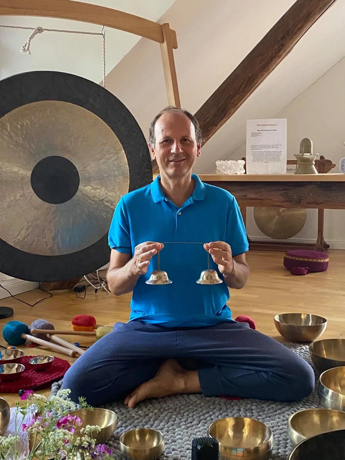Entfalte den Meister in dir! – Meditationen und Klangreisen 4 Remo Rittiner sitzt meditierend vor großem Gong in Meditationsraum, hält zwei Klangschalen-Schlägel, umgeben von Blumen und K
