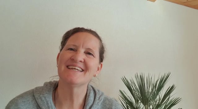 Videostandbild einer Frau mit Dutt, grauem Pullover lächelnd vor weißem Hintergrund