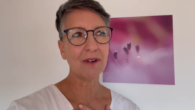 Videostandbild einer Frau mit Brille und hellem Oberteil, lachend vor unscharfem Hintergrund
