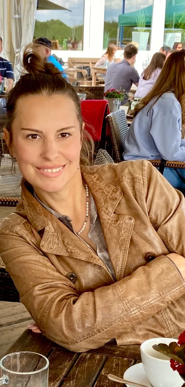 Frau in beiger Jacke mit Perlen-Halskette in Restaurant-Ambiente, lächelnd