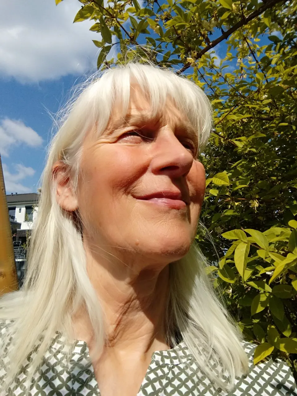 Porträt einer älteren Frau mit weißen Haaren vor grünem Blätterwerk und Himmel