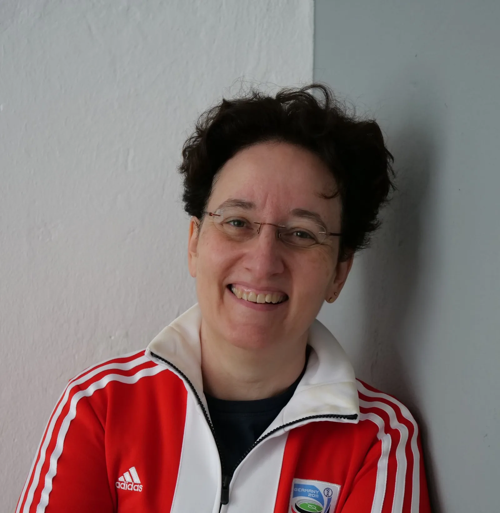 Porträt einer Frau mit Brille in roter Adidas-Jacke, lächelnd vor weißer Wand