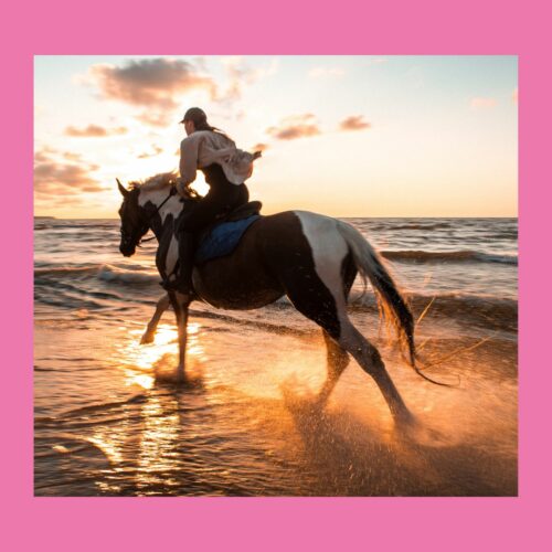 Frau reitet schwarzes Pferd im Sonnenuntergang am Strand entlang