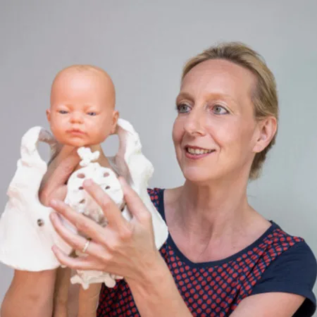 Expedition Familie – Videokurs für werdende Eltern 15 Katharina Jeschke hält lächelnd ein Baby in weißer Kleidung auf neutralem grauem Hintergrund