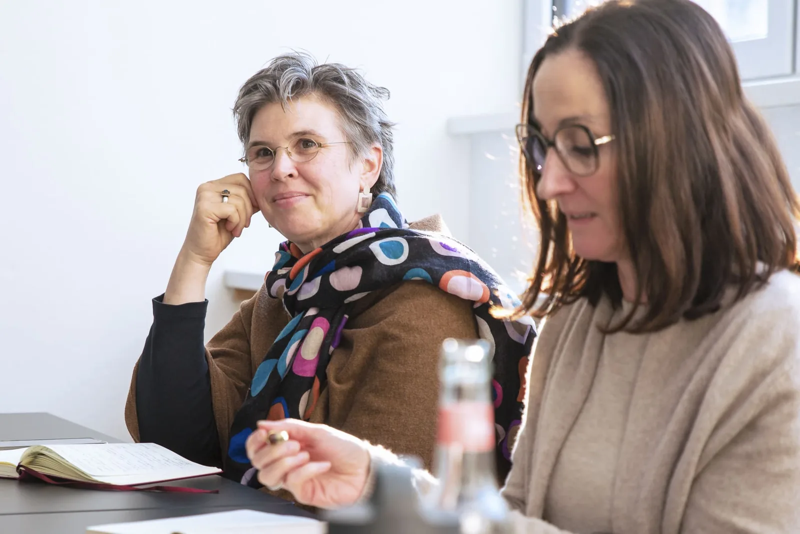 Zwei Frauen an Tisch mit Unterlagen – eine mit braunem Blazer, eine mit Brille