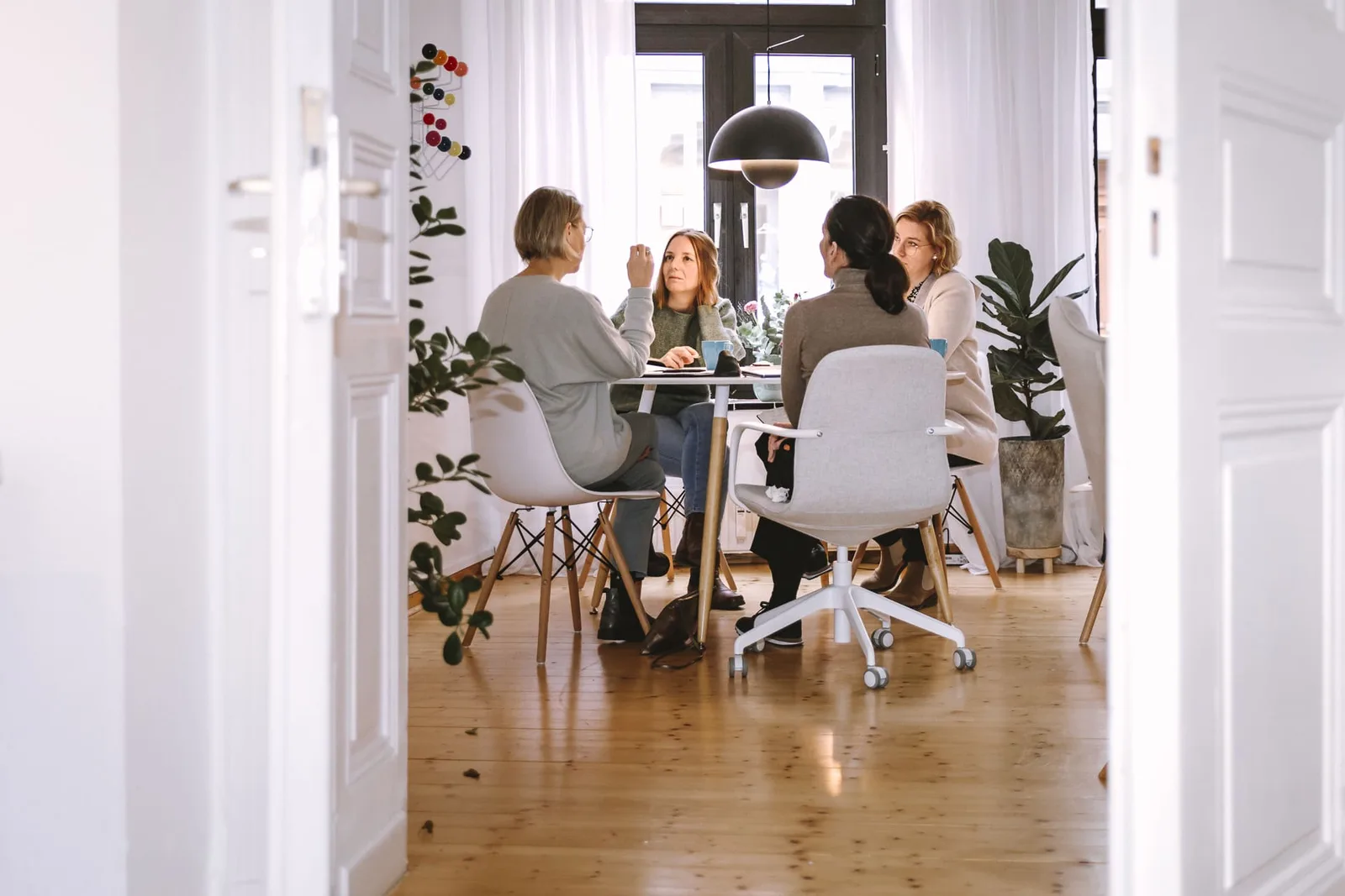 Vier Frauen sitzen an Tisch in hellem Coworking-Space mit Pflanzen und schwarzer Lampe