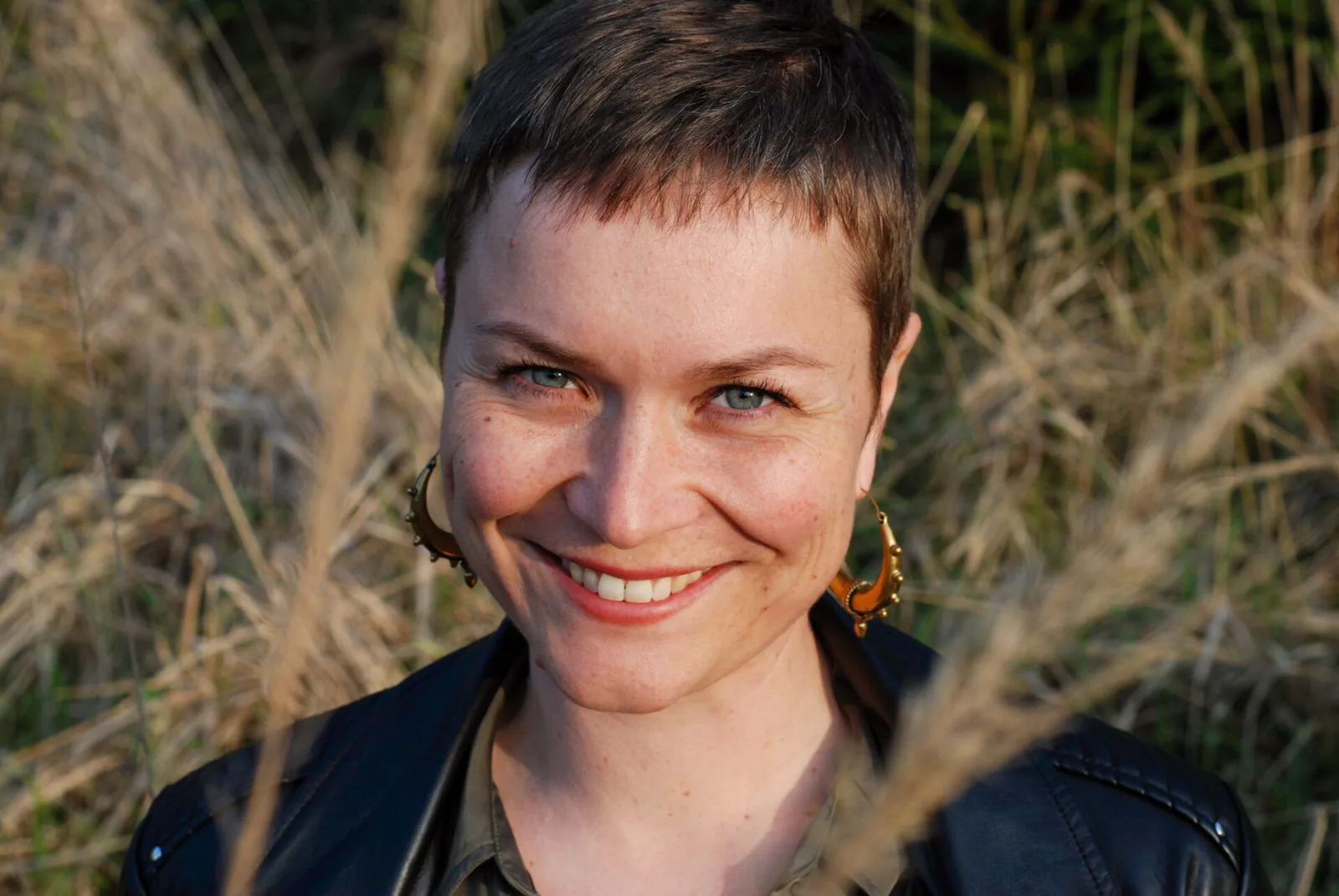 Frau mit kurzen dunklen Haaren und Ohrringen lächelt vor Grasgrund – Anna Buchty riot hearts