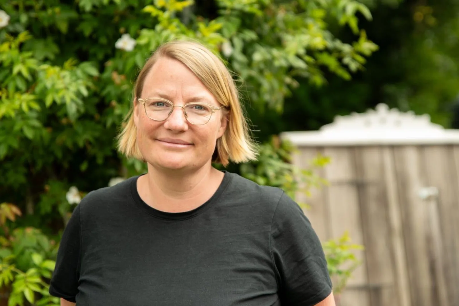 Blonde Frau mit Brille im schwarzen Shirt vor grünem Gartenhintergrund – Eva Wieners Munich Fundraising School