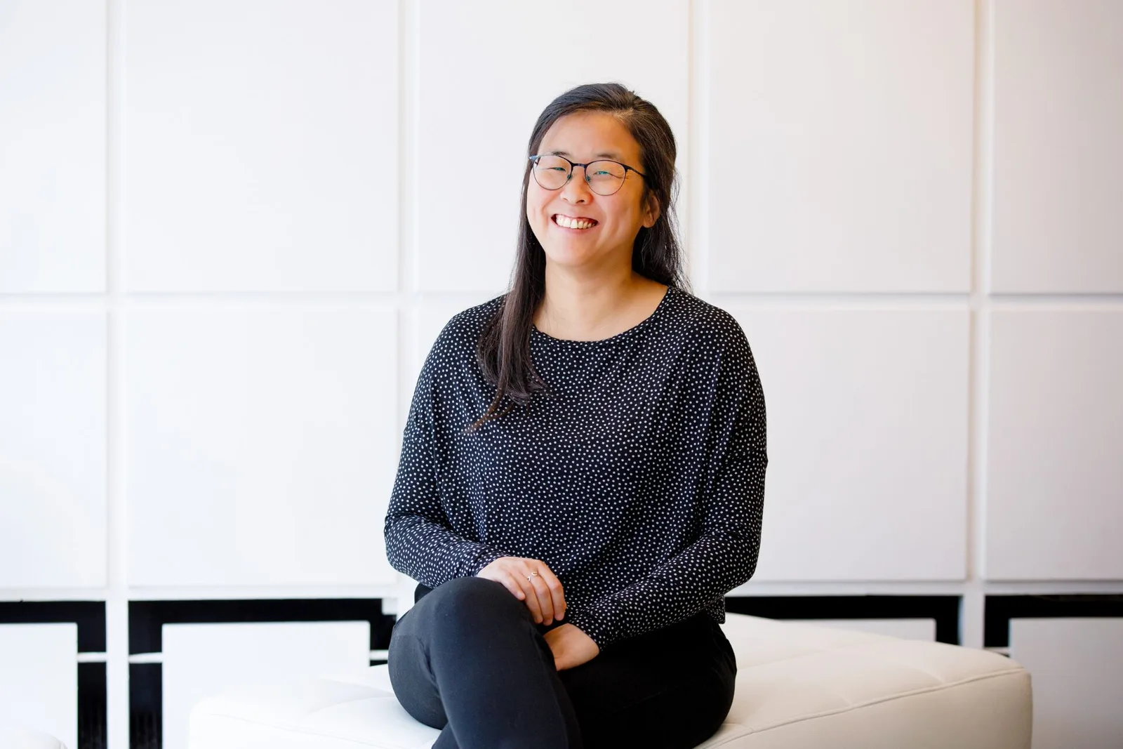 Junge Frau mit Brille und Polka-Dot-Shirt lächelt vor heller Wand – Testimonial Esther Hur step2balance