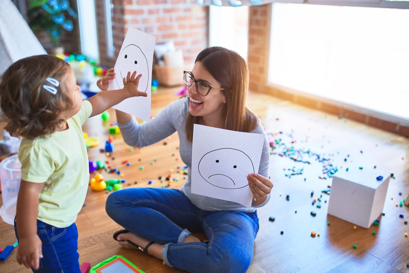 Erzieherin mit Kind hält Papiere mit Emoticon-Gesichtern in hellem Raum