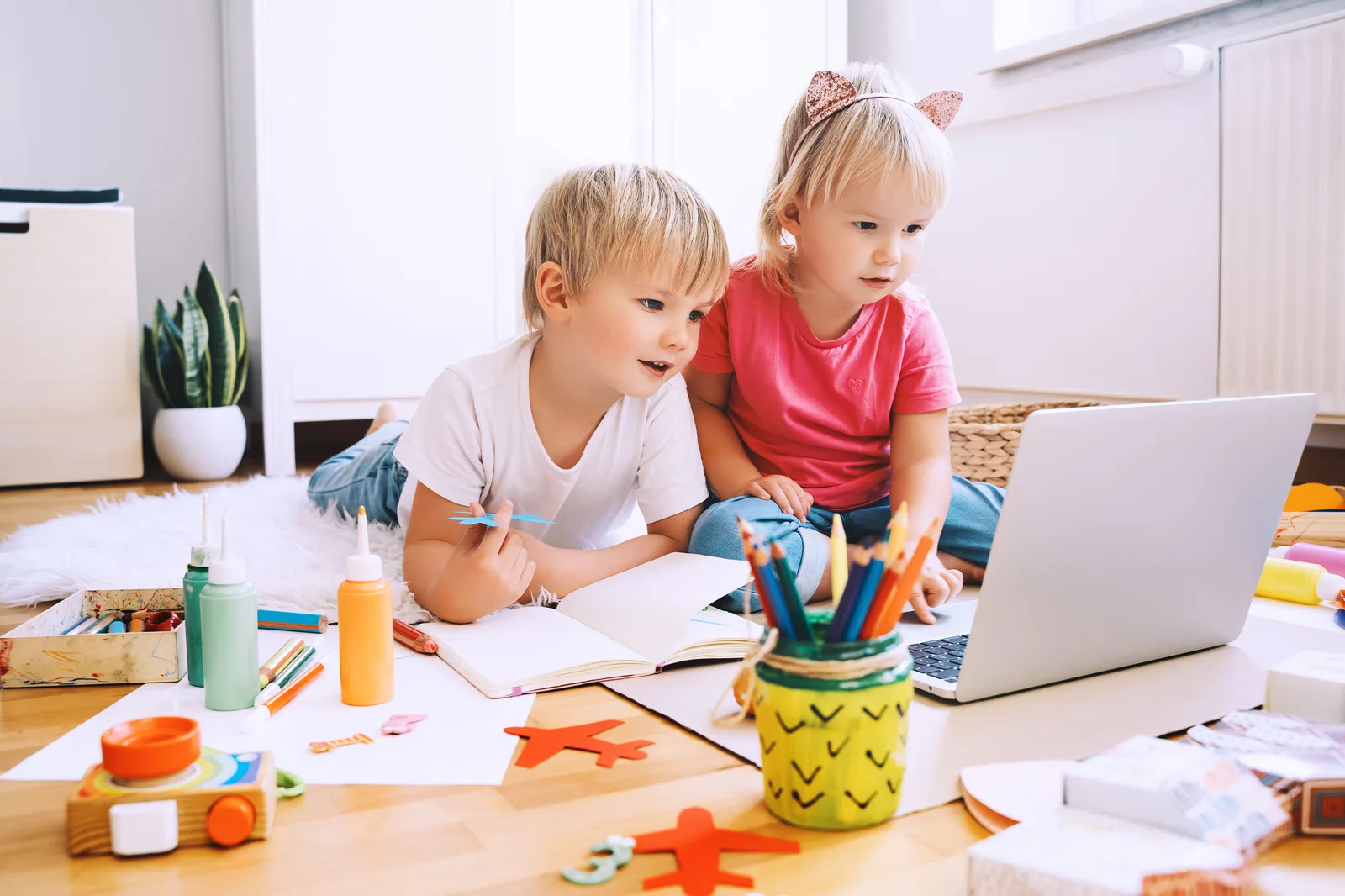 Zwei Kinder arbeiten gemeinsam am Laptop, umgeben von Bastelmaterialien und Stiften