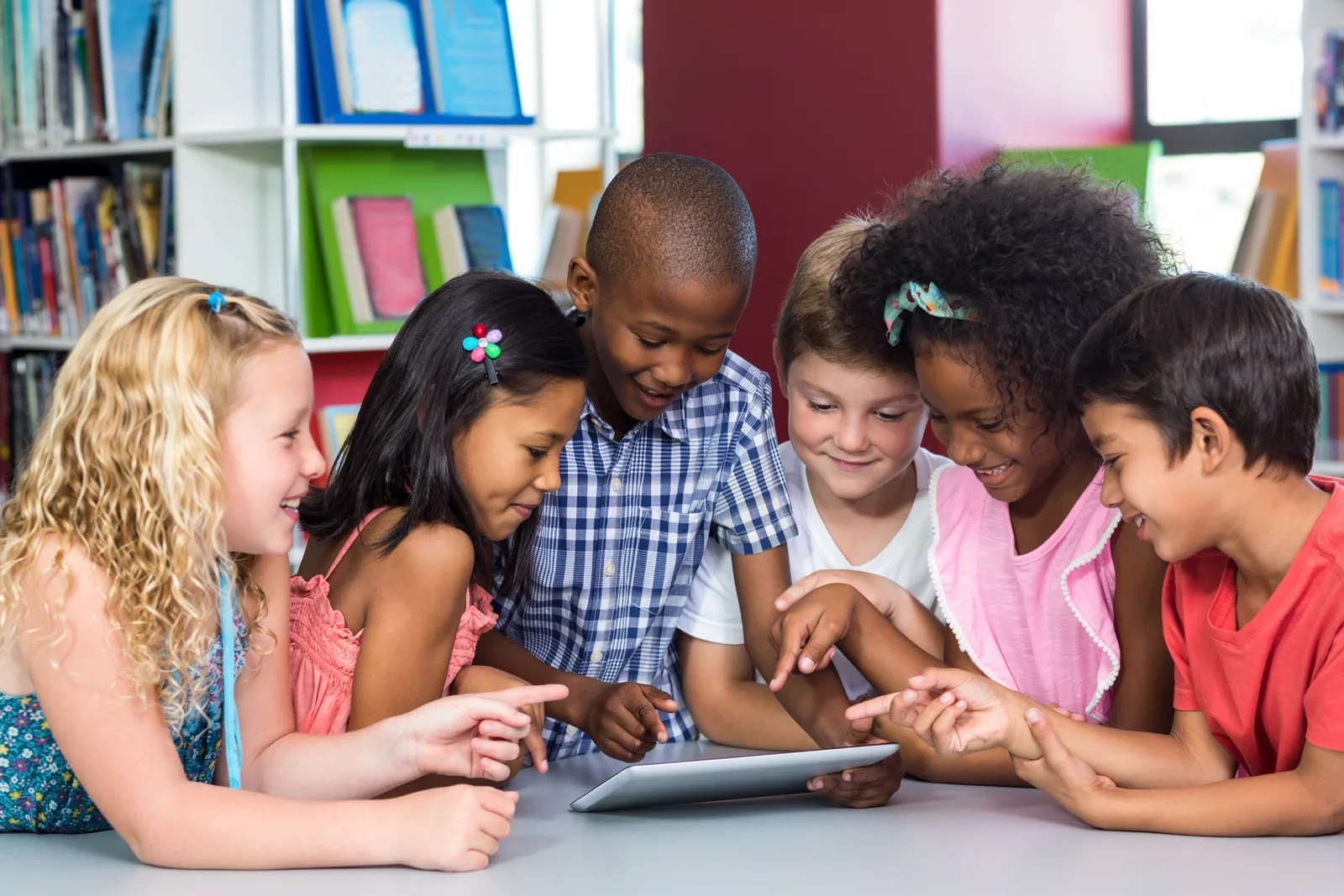Sechs Kinder verschiedener Ethnien sitzen zusammen an einem Tisch und schauen auf ein Tablet, bunte Bücherregale im Hintergru
