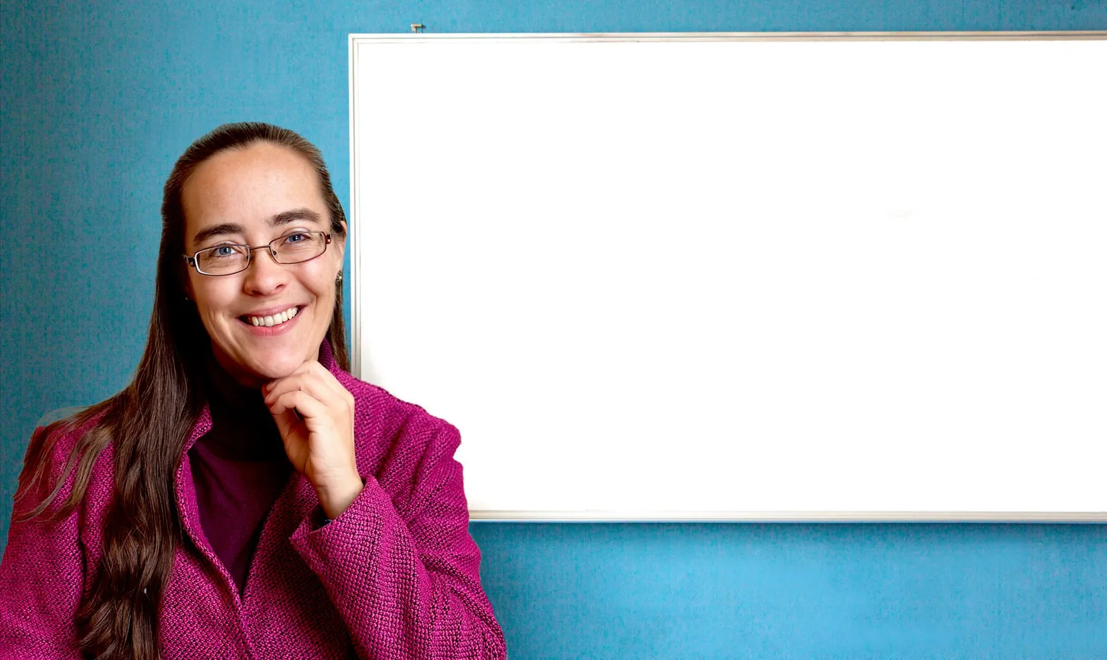 Ricarda Colditz vor türkisblauer Wand neben leerem Whiteboard, lila Wollblazer, Brille, lächelnd