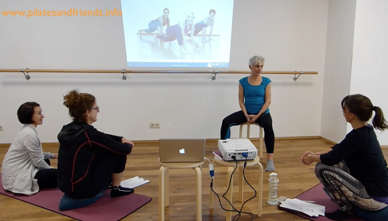 Workshopsituation mit Instructorin auf Stuhl und vier sitzenden Teilnehmern auf Matten