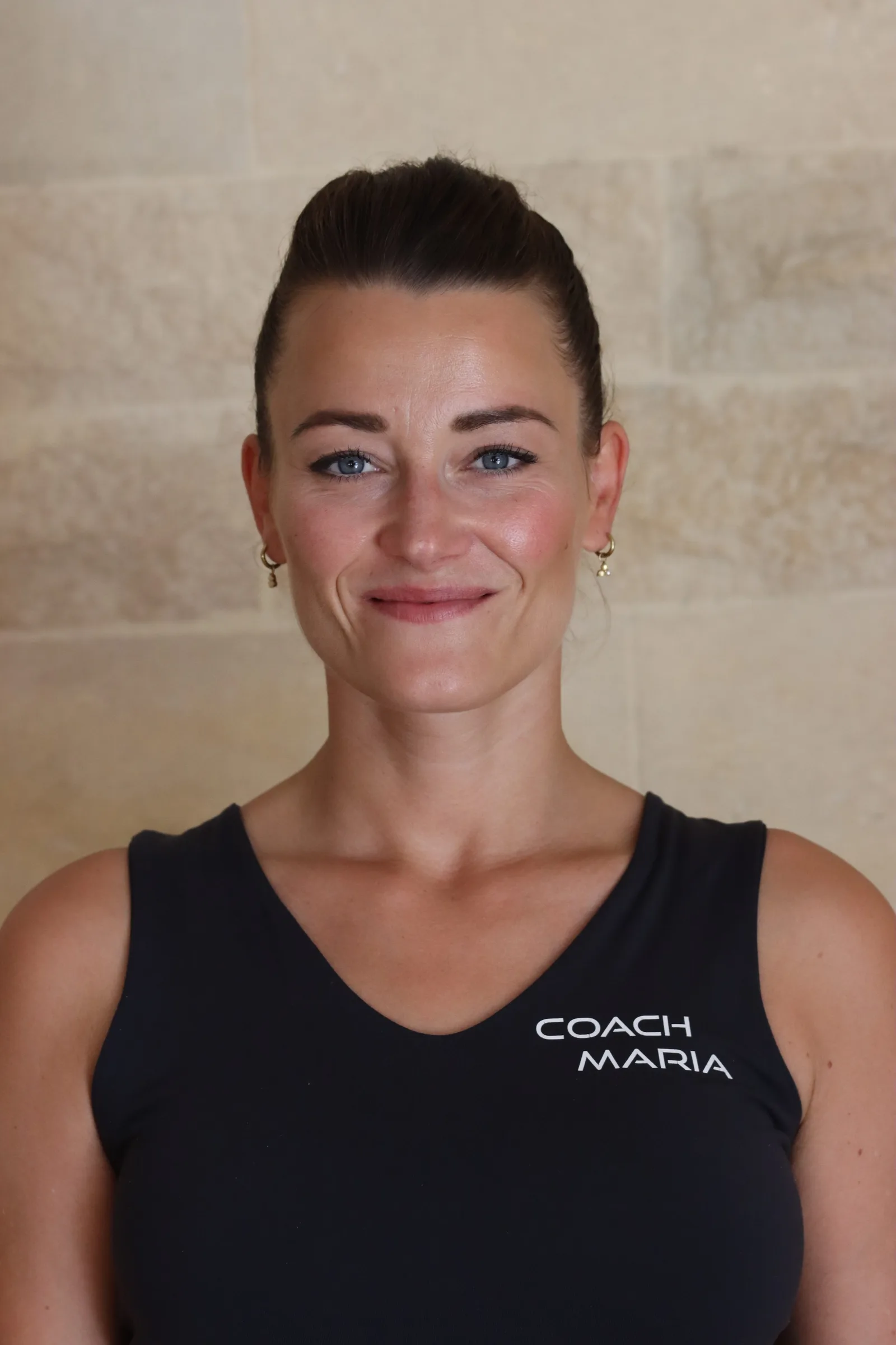 Frau mit dunklem Haar im schwarzen Tanktop mit "COACH MARIA" Schriftzug sitzt frontal vor Steinwand