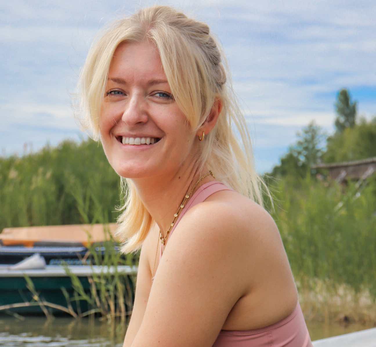 Blonde Frau in Rosa-Top am See lächelnd, Bäume und Wasser im Hintergrund