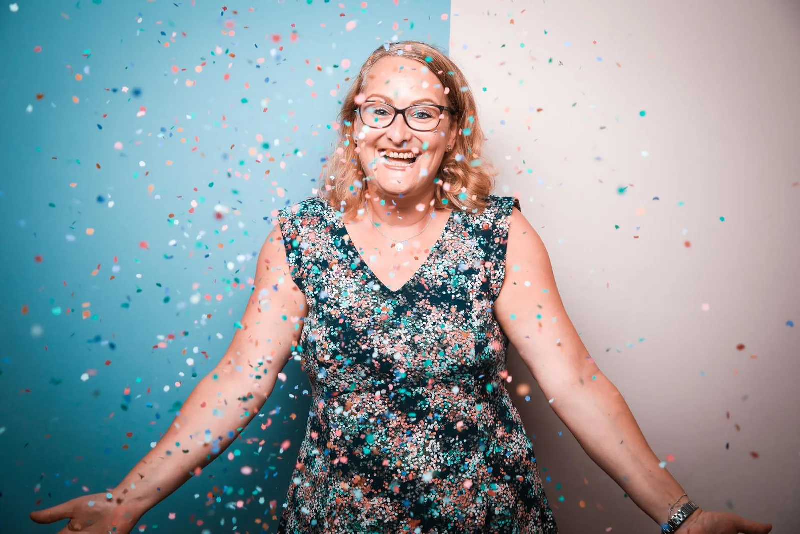 Lachende Frau mit Brille und geblümtem Kleid vor blauem Hintergrund mit buntem Konfetti