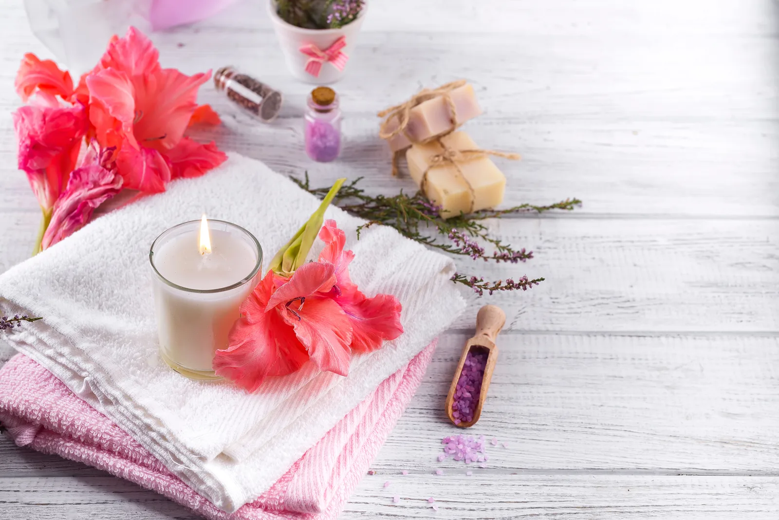 Spa-Arrangement mit rosa Blüten, brennender Kerze, Seife, Badesalz und Handtüchern auf Holzoberfläche