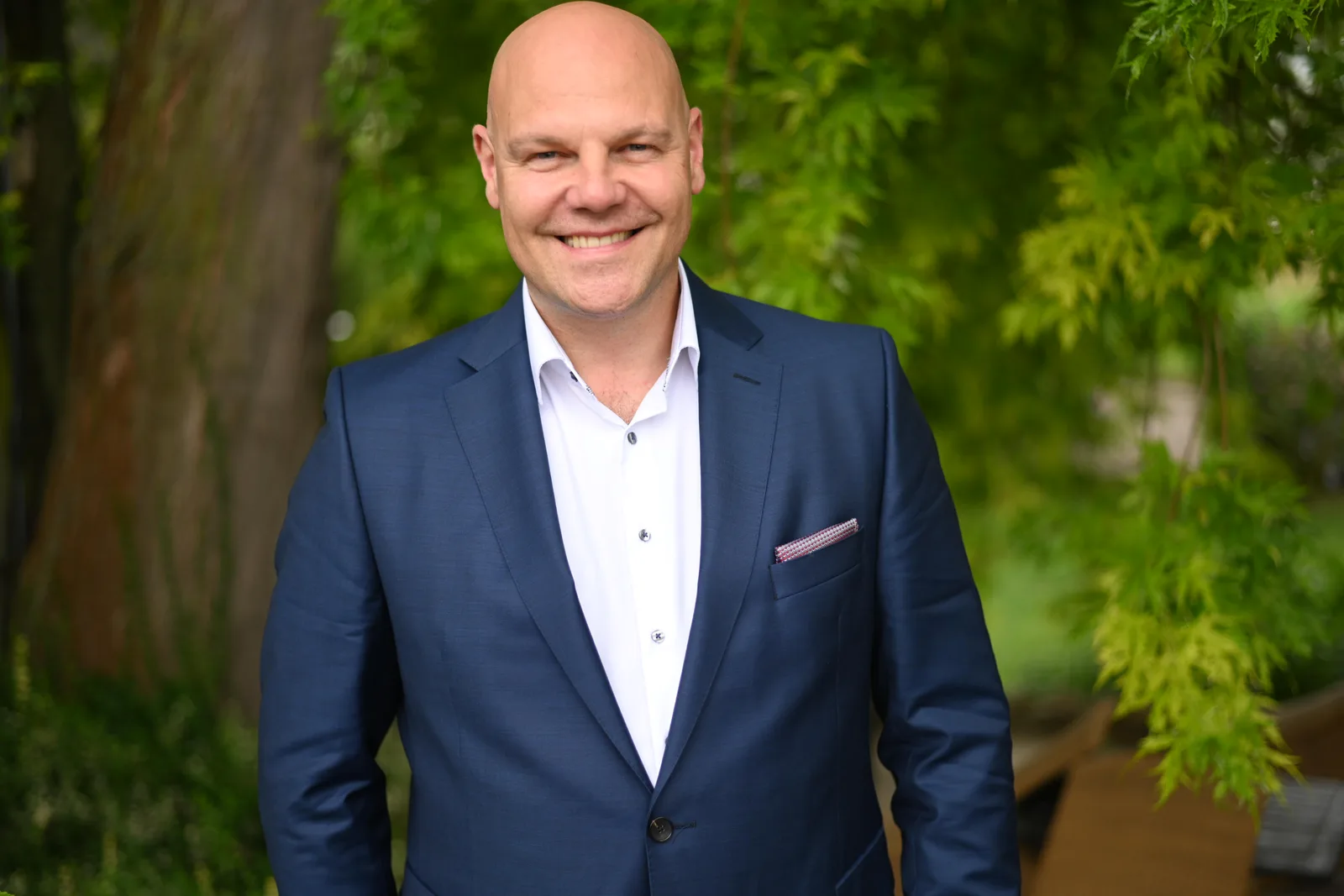 Ingo Steinbock in dunklem Anzug mit weißem Hemd im Freien vor grünem Laub