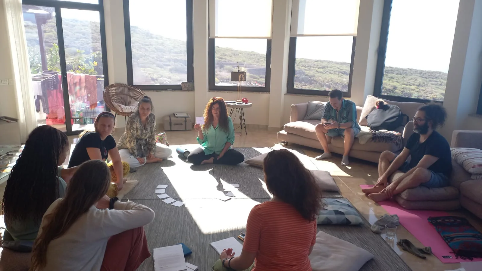 Seminarraum mit großen Fenstern und Bergsicht, Teilnehmer sitzen im Kreis auf Yogamatten, Trainerin sitzt zentral in grünem O