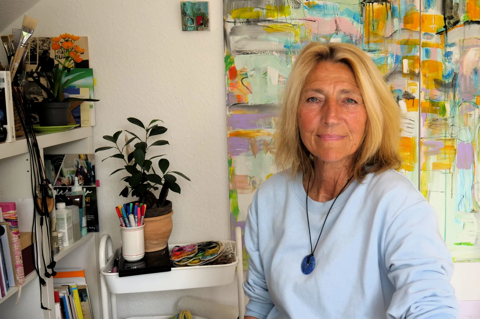 Karin Henningsen sitzt vor farbigem abstraktem Wandbild in hellem Atelier mit Pflanzen