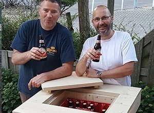 Zwei Männer demonstrieren ein DIY-Projekt mit Holzkonstruktion an einem Tisch im Freien