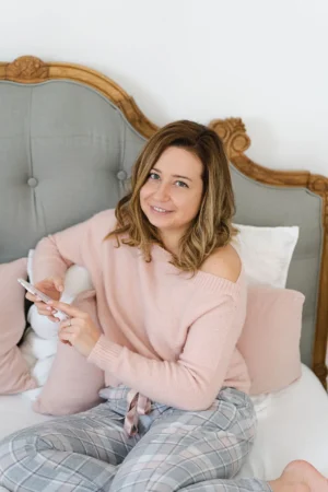 Lilli Koisser sitzt lächelnd auf einem Bett mit grauem Kopfteil, trägt einen rosa Pullover und hält ein Smartphone in den Hän