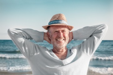 Mann mittleren Alters mit Hut am Strand, entspannt lächelnd, Hände hinter Kopf