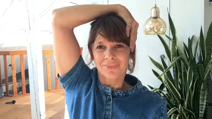 Frau in Jeanshemd mit Hand am Nacken, Blick zur Seite, helles Wohnzimmer mit Pflanze