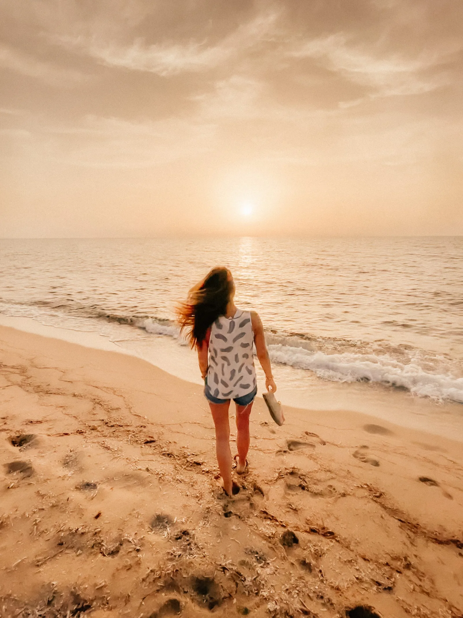 Frau mit rötlichen Haaren läuft bei Sonnenuntergang am Strand entlang, trägt weißes Muster-Top und hält Schuhe in der Hand