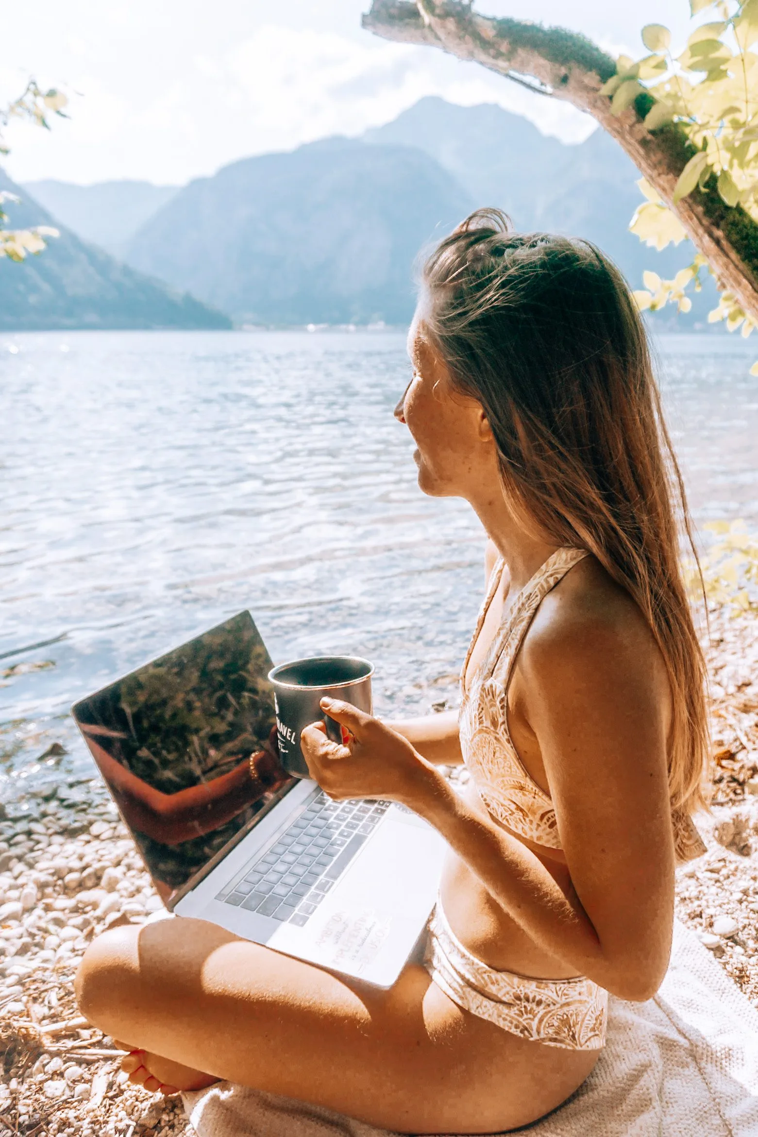 Frau sitzt am Seeufer mit Laptop und Kaffeetasse, Berglandschaft im Hintergrund
