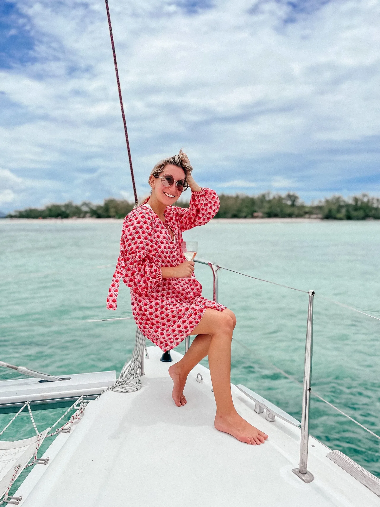 Frau in rotem Muster-Hemdkleid auf einem Segelboot (Katamaran), lächelnd mit Sonnenbrille