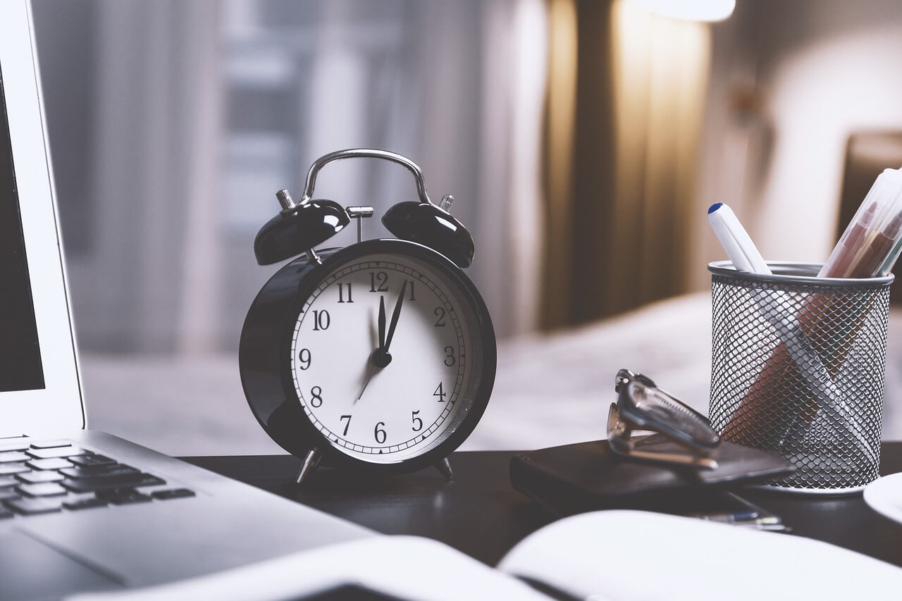 Schwarze Wecker-Uhr auf Bürotisch neben Laptop, Stifthalter und Papieren, symbolisiert Zeitmanagement