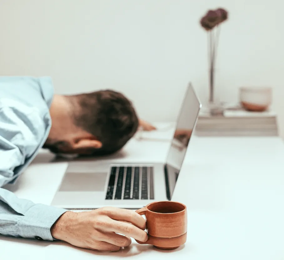 Erschöpfter Mann mit Kopf auf Laptop liegend am Schreibtisch mit Kaffeebecher, symbolisiert Überbelastung