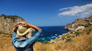 Frau mit Strohhut blickt auf Mittelmeerküste mit Felsen und blauem Meer